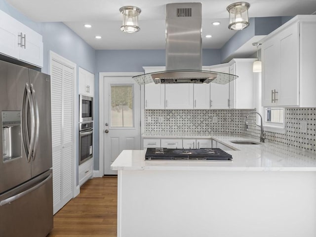 kitchen with island exhaust hood, appliances with stainless steel finishes, white cabinets, a sink, and a peninsula