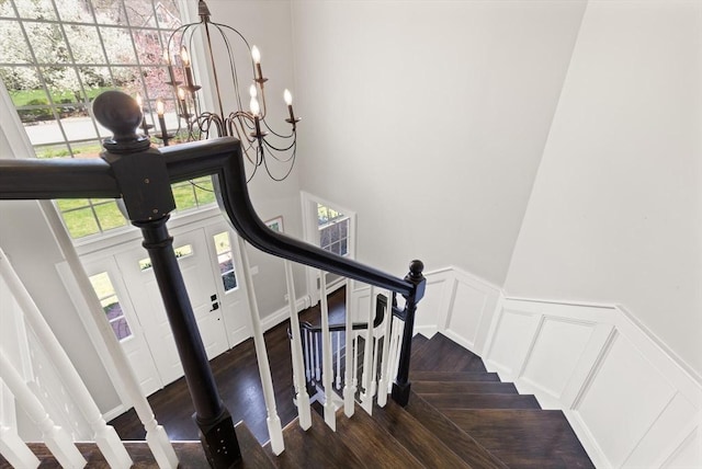 stairway with a towering ceiling, wainscoting, wood finished floors, an inviting chandelier, and a decorative wall