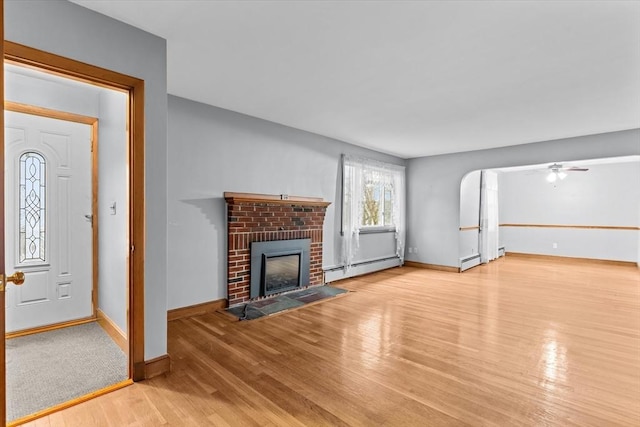 unfurnished living room with arched walkways, a baseboard heating unit, wood finished floors, baseboards, and a brick fireplace