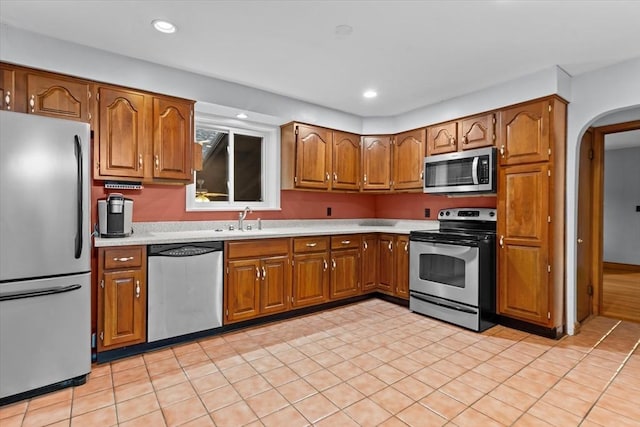 kitchen featuring arched walkways, appliances with stainless steel finishes, brown cabinets, light countertops, and a sink