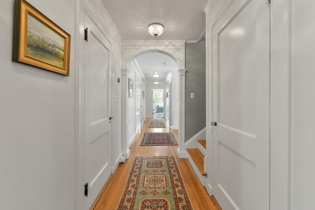 hallway with light wood-type flooring, crown molding, ornate columns, a decorative wall, and arched walkways