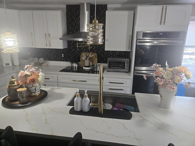 kitchen featuring light stone countertops, stainless steel double oven, wall chimney range hood, backsplash, and white cabinets