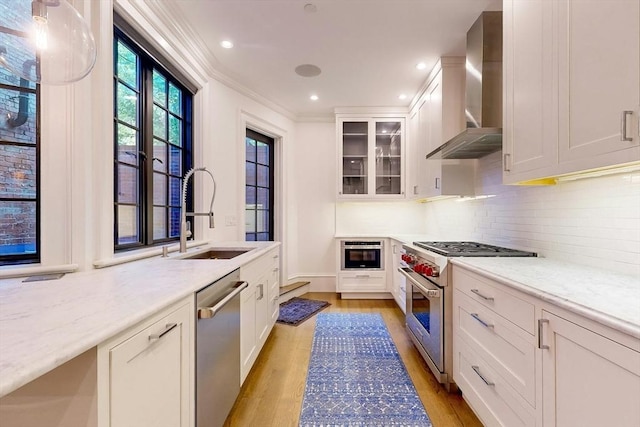 kitchen with light stone counters, stainless steel appliances, sink, wall chimney range hood, and white cabinets