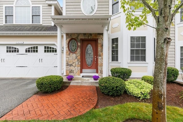 property entrance featuring an attached garage, asphalt driveway, roof with shingles, and stone siding