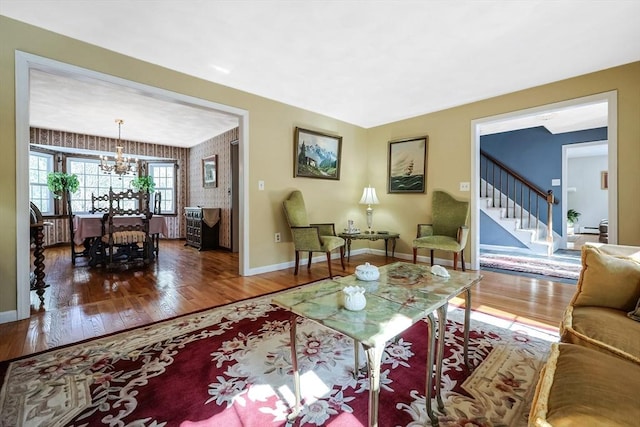 living room featuring a notable chandelier, stairway, wood finished floors, baseboards, and wallpapered walls