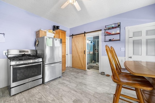 kitchen featuring a textured ceiling, ceiling fan, a barn door, appliances with stainless steel finishes, and wainscoting