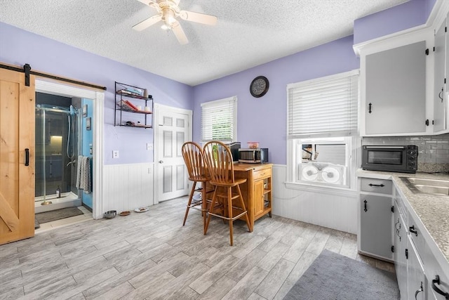 office area with a barn door, a ceiling fan, wainscoting, light wood-style flooring, and a textured ceiling