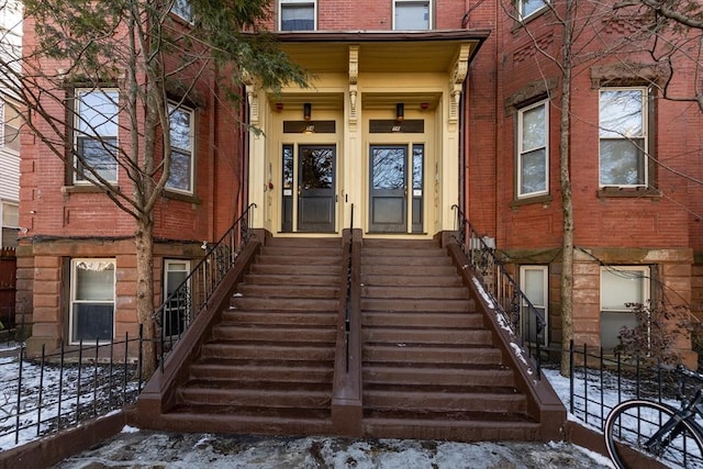 snow covered property entrance featuring french doors, brick siding, and fence