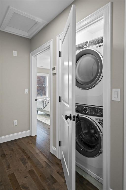 clothes washing area featuring stacked washer and dryer, attic access, laundry area, baseboards, and dark wood-style flooring