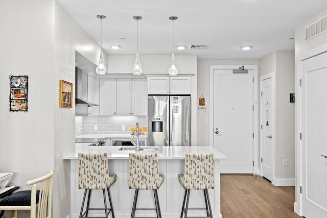 kitchen featuring stainless steel refrigerator with ice dispenser, wall chimney exhaust hood, a peninsula, and light countertops