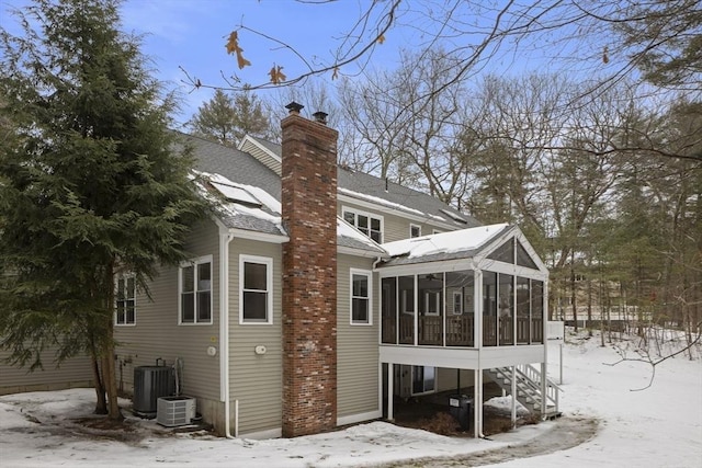 snow covered rear of property featuring a sunroom, a shingled roof, a chimney, and central AC unit