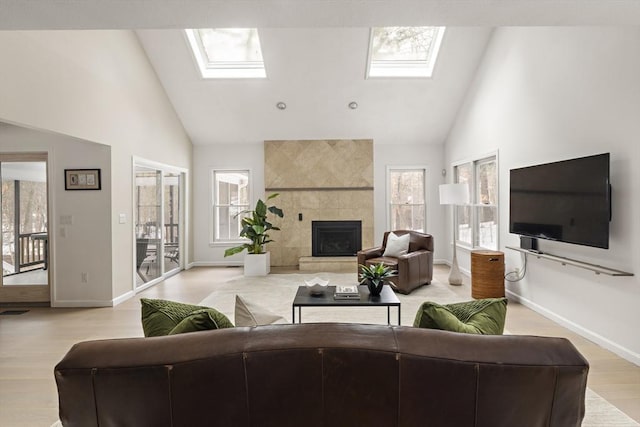 living area featuring a skylight, light wood finished floors, baseboards, a tiled fireplace, and high vaulted ceiling