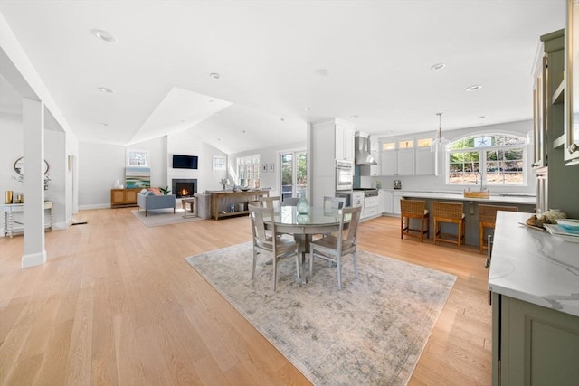 dining room with light wood finished floors, a healthy amount of sunlight, a lit fireplace, and vaulted ceiling