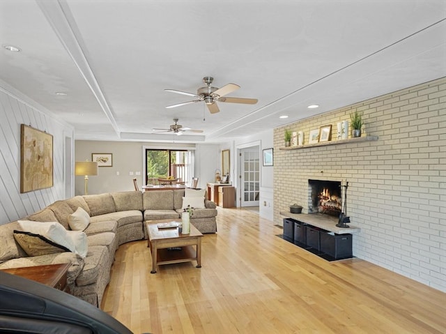 living area featuring ceiling fan, brick wall, a fireplace, light wood-type flooring, and a raised ceiling