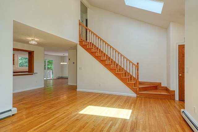 unfurnished living room featuring a baseboard heating unit, hardwood / wood-style flooring, stairs, a towering ceiling, and a skylight