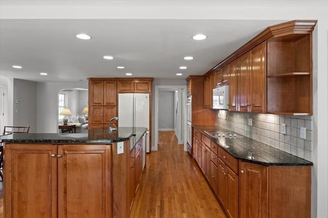 kitchen featuring brown cabinetry, open shelves, white appliances, an island with sink, and arched walkways