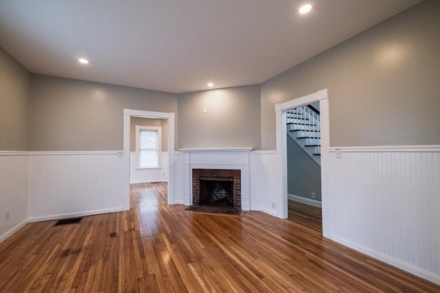 unfurnished living room featuring a wainscoted wall, stairs, wood finished floors, a brick fireplace, and recessed lighting
