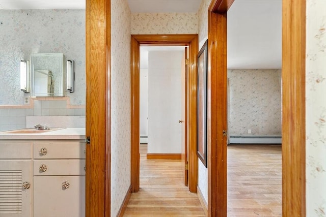 corridor with a baseboard heating unit, a sink, baseboard heating, light wood-type flooring, and wallpapered walls