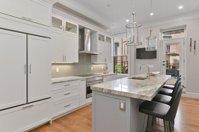 kitchen with wall chimney range hood, white cabinetry, sink, and a kitchen island with sink