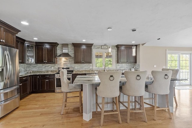 kitchen featuring stainless steel appliances, wall chimney exhaust hood, a breakfast bar, backsplash, and dark brown cabinets