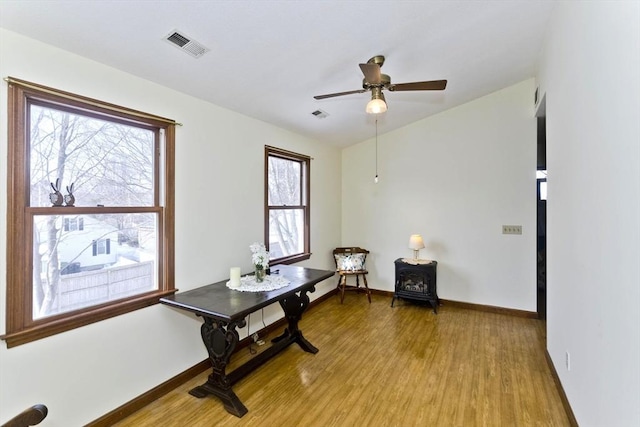 office space featuring light wood finished floors, visible vents, a ceiling fan, a wood stove, and baseboards