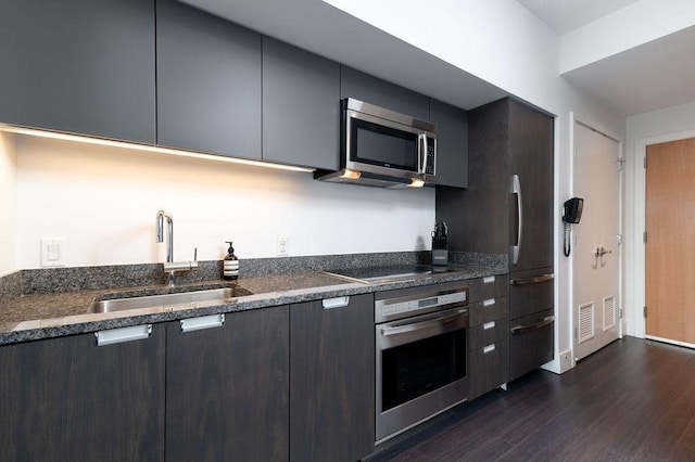 kitchen with appliances with stainless steel finishes, a sink, dark wood-style floors, modern cabinets, and dark stone countertops