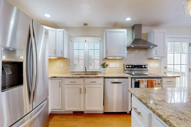 kitchen featuring light wood-style flooring, appliances with stainless steel finishes, wall chimney range hood, white cabinetry, and a sink