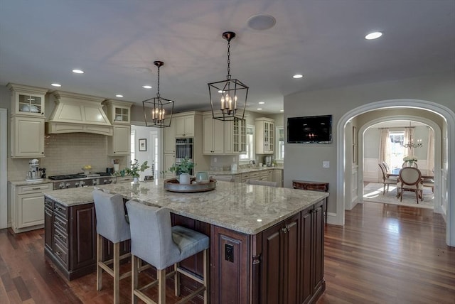 kitchen featuring a breakfast bar area, premium range hood, light stone countertops, decorative light fixtures, and a large island with sink