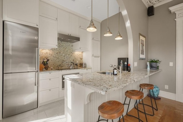 kitchen with a sink, a peninsula, under cabinet range hood, light stone countertops, and appliances with stainless steel finishes