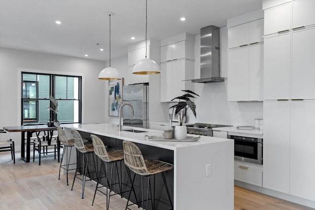 kitchen featuring wall oven, a kitchen island with sink, pendant lighting, stainless steel refrigerator, and wall chimney exhaust hood