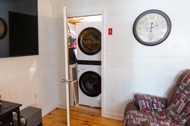 clothes washing area featuring light wood-type flooring, stacked washing maching and dryer, laundry area, and a wainscoted wall