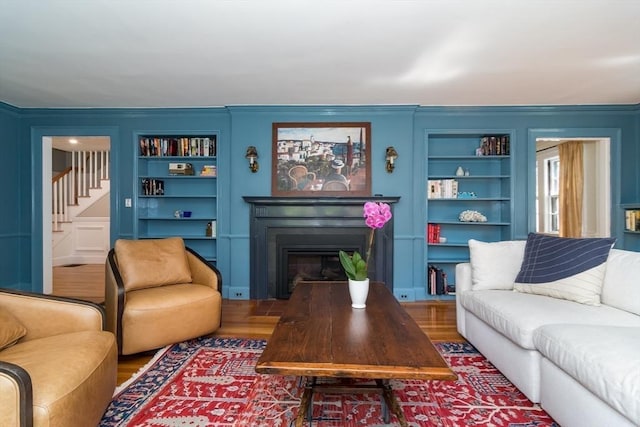 living room featuring a fireplace, built in shelves, crown molding, stairs, and wood finished floors