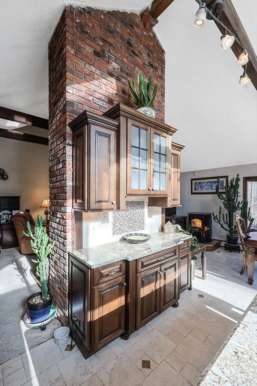 bar featuring backsplash, light stone counters, dark brown cabinets, vaulted ceiling with beams, and a wood stove