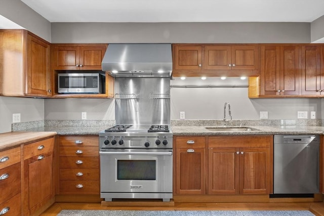 kitchen with appliances with stainless steel finishes, brown cabinets, light stone countertops, wall chimney range hood, and a sink