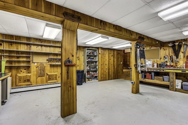 basement with a baseboard heating unit, a drop ceiling, wood walls, stairway, and a workshop area