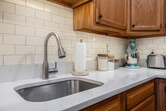 kitchen featuring decorative backsplash, light stone counters, brown cabinetry, and a sink