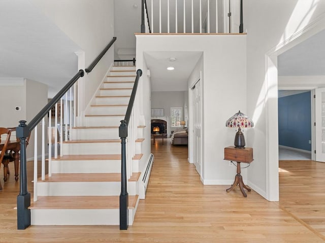 stairway with crown molding, a baseboard heating unit, baseboards, a lit fireplace, and wood finished floors