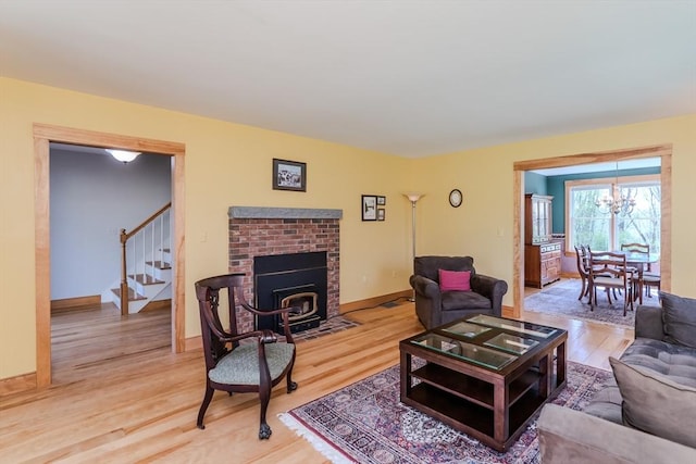 living room featuring an inviting chandelier, stairs, a brick fireplace, light wood-style floors, and baseboards