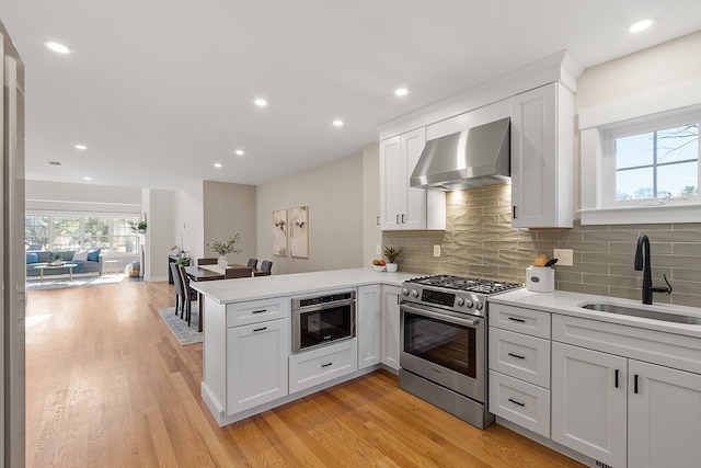 kitchen featuring stainless steel appliances, light wood-type flooring, sink, kitchen peninsula, and wall chimney exhaust hood