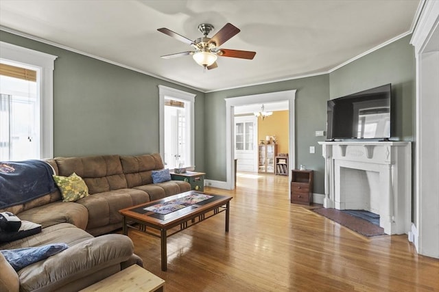 living area with crown molding, wood finished floors, baseboards, a fireplace with flush hearth, and ceiling fan with notable chandelier