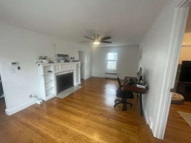 living area featuring a ceiling fan, a fireplace with flush hearth, radiator heating unit, light wood-type flooring, and baseboards