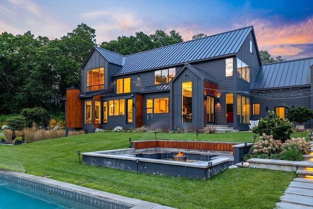 back of property at dusk with an outdoor fire pit, metal roof, board and batten siding, a yard, and a standing seam roof