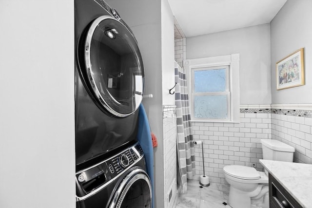 bathroom with toilet, vanity, stacked washer / drying machine, marble finish floor, and wainscoting