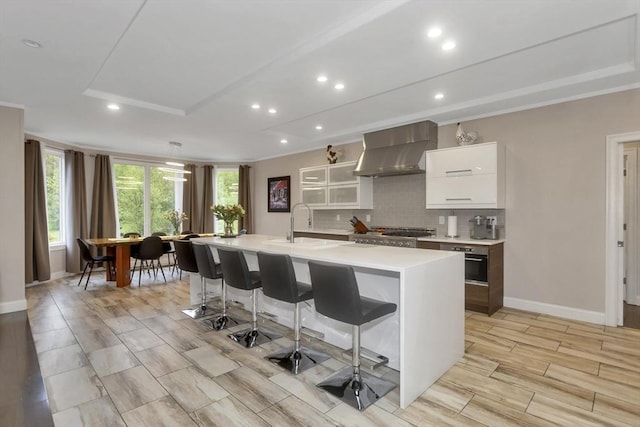 kitchen with wall chimney exhaust hood, light countertops, a breakfast bar area, white cabinetry, and an island with sink