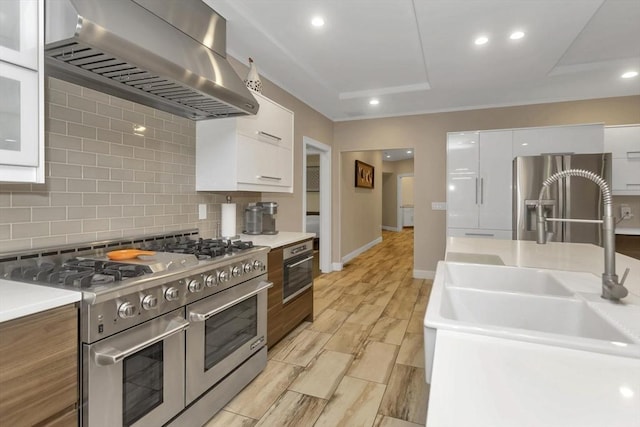 kitchen with stainless steel appliances, wall chimney range hood, a sink, white cabinets, and recessed lighting
