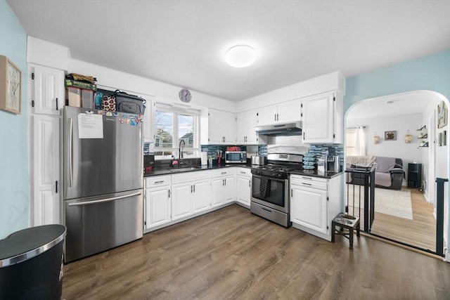 kitchen featuring arched walkways, stainless steel appliances, dark countertops, a sink, and under cabinet range hood