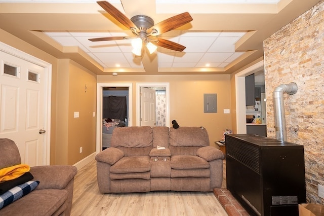 living room featuring light wood-type flooring, ceiling fan, a wood stove, electric panel, and a tray ceiling