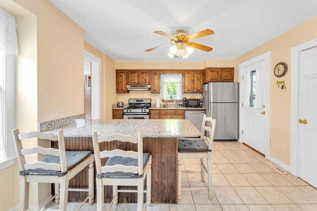 252 Railroad Ave, Norwood, MA 02062 - kitchen with appliances with stainless steel finishes, under cabinet range hood, brown cabinetry, a kitchen bar, and a ceiling fan