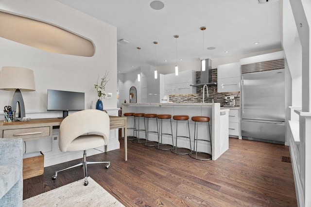 kitchen with built in refrigerator, wall chimney range hood, modern cabinets, decorative backsplash, and white cabinets