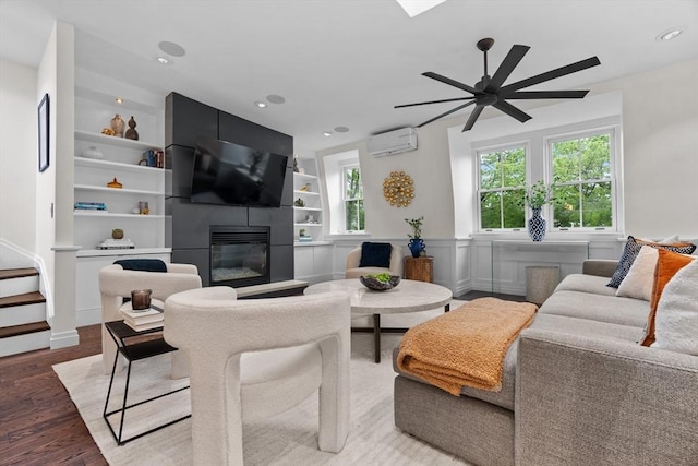 living room with an AC wall unit, wood finished floors, a large fireplace, a ceiling fan, and built in features